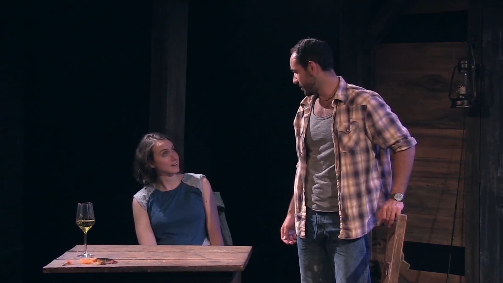 Two actors share a scene at a rustic table on stage