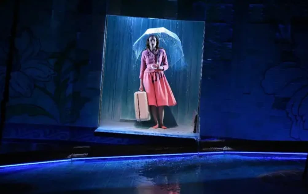 Actress with suitcase and umbrella stands in stage doorway, rain backdrop