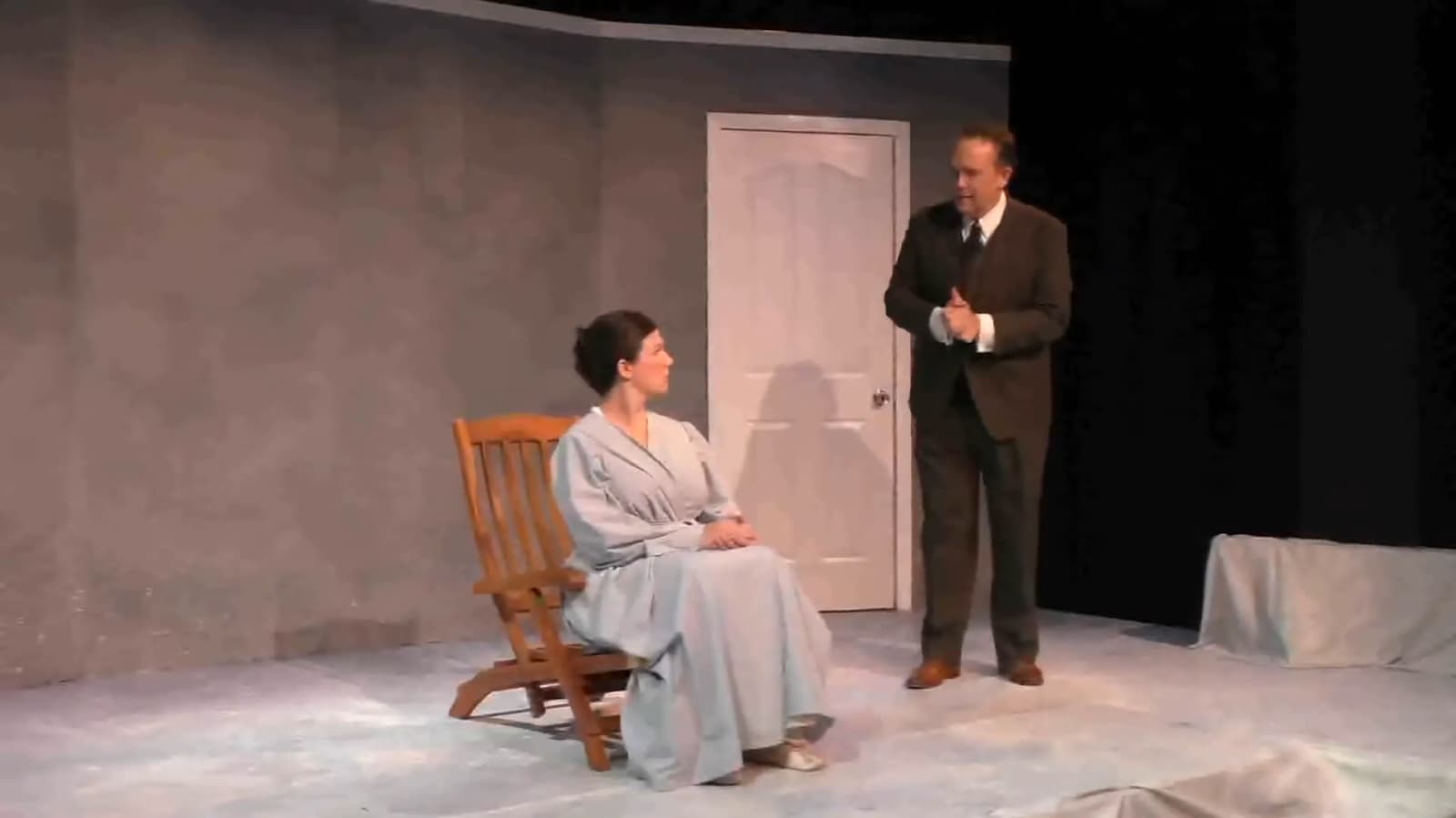 A man in a suit stands talking to a seated woman in a stage play setting