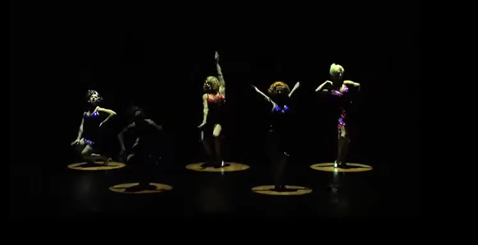 Five performers in sparkling costumes dancing under spotlights