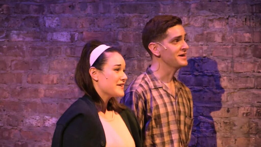 Two actors on stage, smiling with a brick wall backdrop