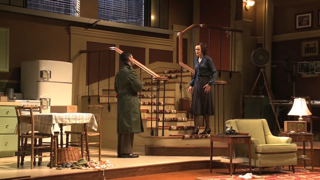A stage set depicts a vintage interior with two actors in a tense scene