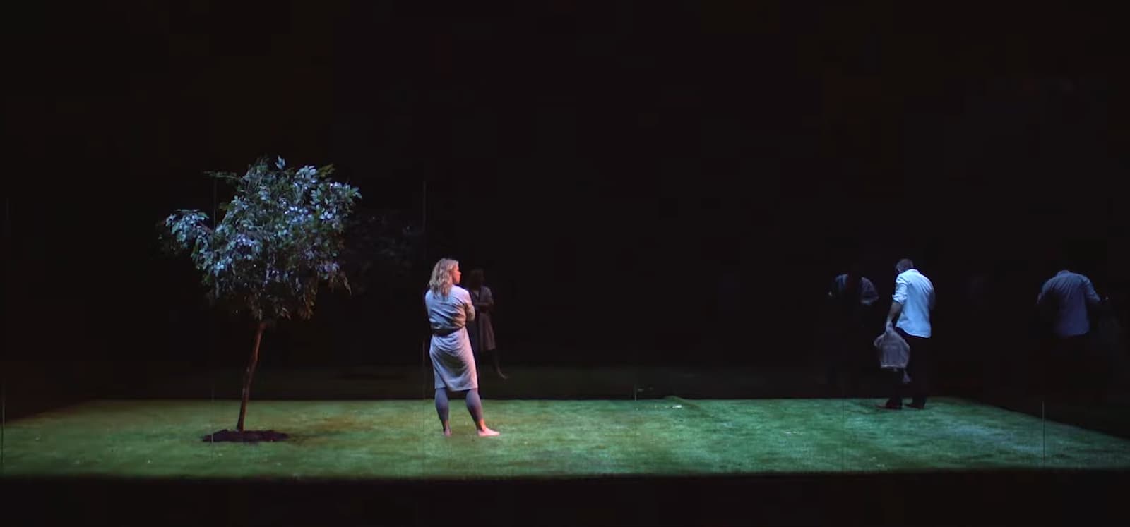 A stage with a single tree under a spotlight, people in the background