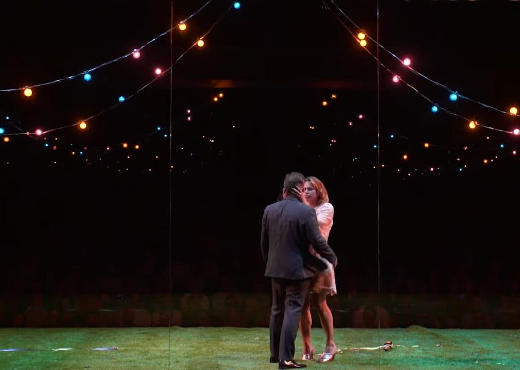 Two people embrace under strings of colorful lights on a stage