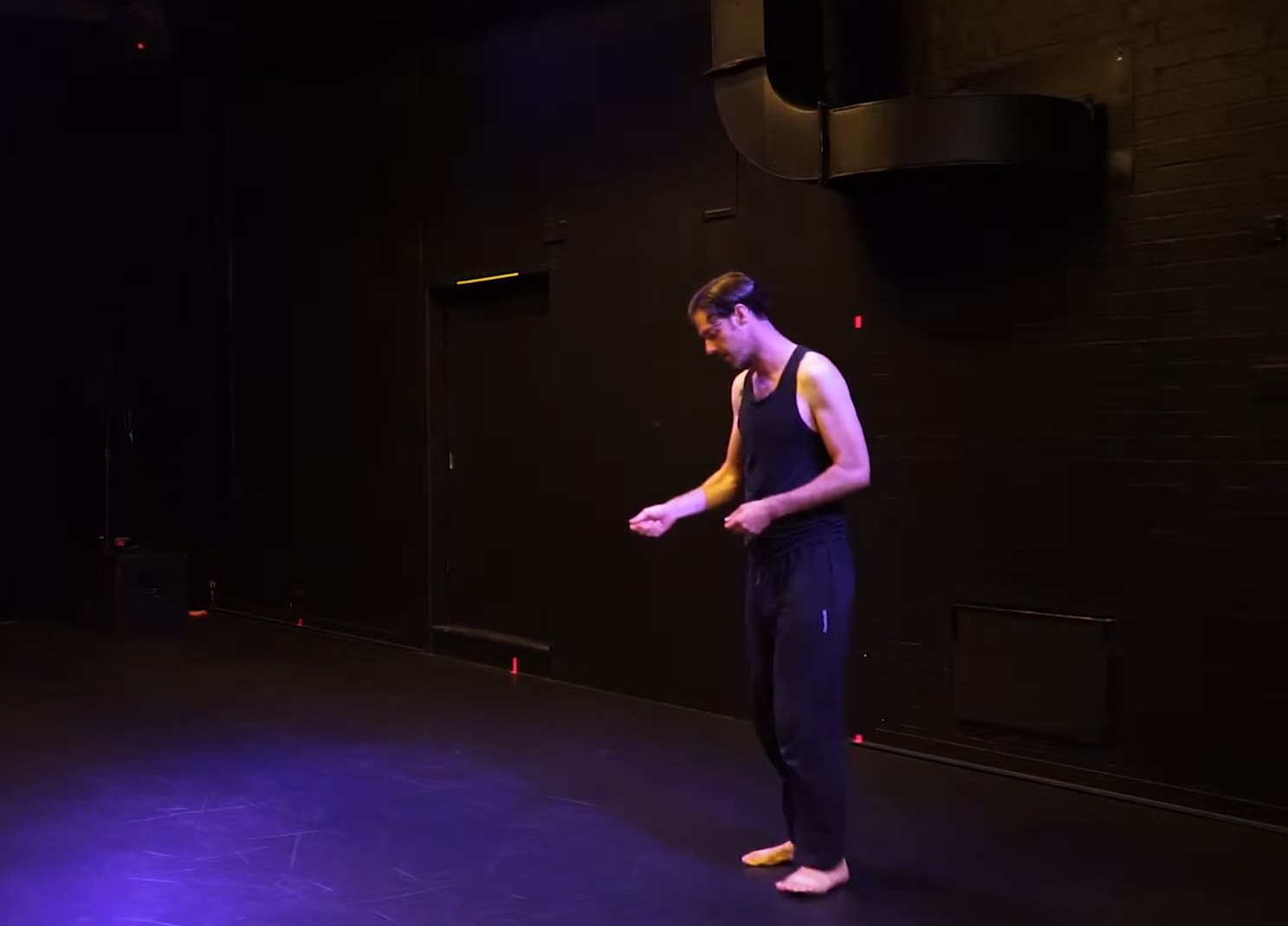 A man in a black tank top and sweatpants checks his wristwatch on a theater stage